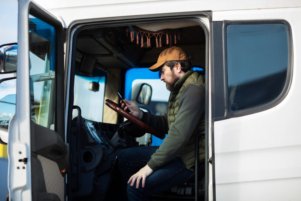 A truck driver sitting in his truck, looking at his smartphone. He is wearing a cap and a vest.